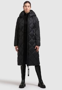 Cămașă puffer neagră, matlasată, cu glugă, design lung, cu fermoar frontal și buzunare laterale. Are un etichetă din material cu numele brandului lângă fermoar.