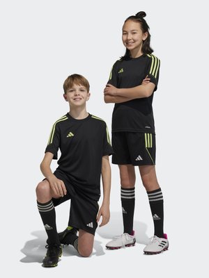 Ropa deportiva negra con franjas de color amarillo neón, combinada con calcetas negras y tacos de fútbol. Dos niños posando, uno de ellos arrodillado.