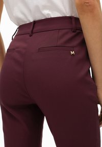 Pantalones de corte burgundy con una textura suave, que cuentan con dos bolsillos traseros y un acento dorado en forma de "M" en el bolsillo derecho.