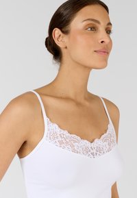 Witte hemdtop met verstelbare banden, voorzien van een bloemenkantafwerking bij de halslijn. Glad, rekbaar materiaal met een aansluitende pasvorm.