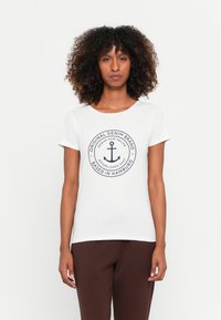 T-shirt branca com decote redondo, mangas curtas e um gráfico central de âncora azul-marinho rodeado de texto, combinada com calças castanhas.