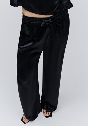 Žmogus dėvi laisvus juodus satino kelnes su elastinga juosmens juosta ir raišteliu, derinamus su juodais atvirų pirštų, kvadratinio kulno sandalais.