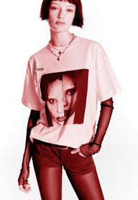 Mladý človek s krátkymi tmavými vlasmi, ktorý má na sebe grafické oversized tričko, vrstvené priesvitné rukavice, kraťasy a náhrdelník, pózuje s rukou v vrecku.