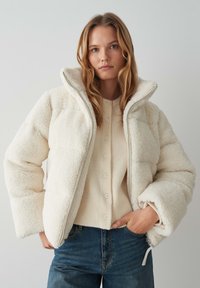 Giacca bianca piumino con un'esterno in tessuto texturizzato simile alla pelliccia, colletto alto e una zip frontale. Indossata sopra un top in maglia color crema.
