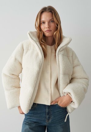 Weiße, puffige Jacke mit einer strukturierten, faux-Shearling-Oberfläche, hohem Kragen und einem Reißverschluss vorne. Getragen über einem cremefarbenen Strickoberteil.