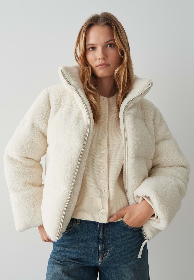Giacca bianca piumino con un'esterno in tessuto texturizzato simile alla pelliccia, colletto alto e una zip frontale. Indossata sopra un top in maglia color crema.