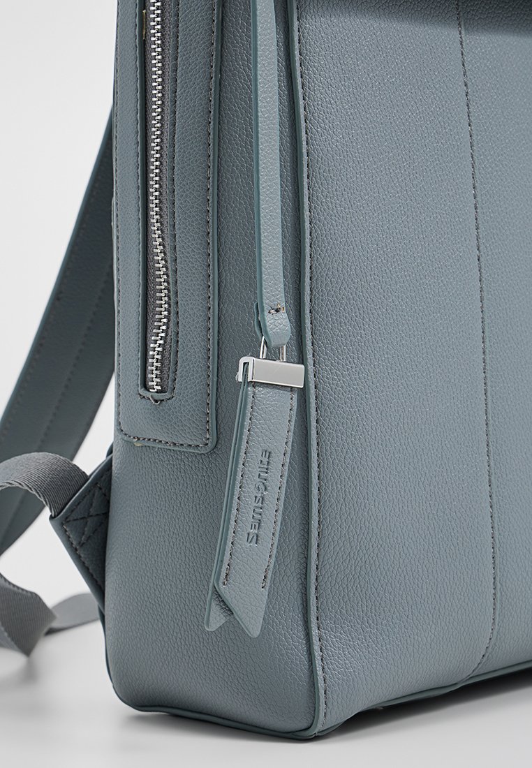 Grau-lederne Rucksack mit strukturierter Oberfläche, ausgestattet mit einem senkrechten Reißverschluss und einem Etikett mit Logo. Verstellbare Stoffgurte sichtbar.