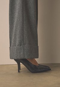Jambe en pantalon gris large avec revers, portant des chaussures à talons hauts noirs à bout pointu sur un sol et un mur beige.
