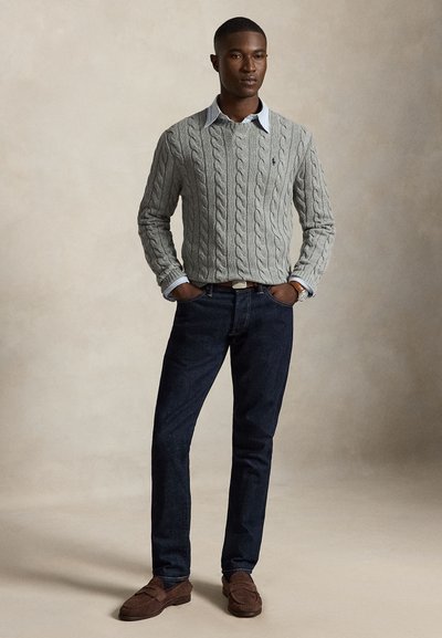 Uomo in piedi con le mani nelle tasche che indossa un maglione grigio lavorato a trecce, una camicia celeste con collo, jeans scuri e mocassini marroni.