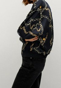 Chaqueta bomber negra con un patrón de cadenas doradas, que presenta una textura suave, puños y cintura acanalados. Tejido brillante con un ajuste relajado.