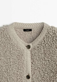 Cardigan beige texturé avec col en V, présentant un design en tricot, deux boutons et une surface duveteuse. Étiquette visible au niveau de l'encolure.