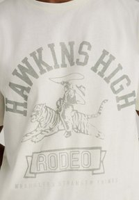 Biały bawełniany t-shirt z szarą grafiką kowboja na tygrysie, z tekstem "HAWKINS HIGH" i "RODEO" w pogrubionej czcionce szeryfowej.