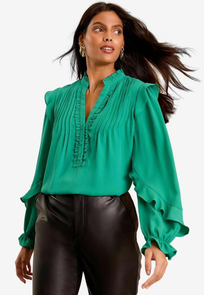 Blusa verde con dettagli arricciati, design plissettato e scollo con bottoni. Maniche lunghe con polsini arricciati. Abbinata a pantaloni in pelle nera.