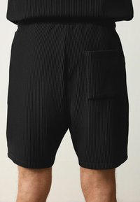 Shorts noirs côtelés avec une taille élastique, dotés d'une seule poche à patch sur la jambe droite. Le tissu texturé est clairement visible.