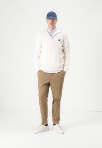 Witte kabelgebreide pullover met rits, lichtblauw overhemd eronder, beige broek en witte sneakers. Blauwe pet gedragen op het hoofd.