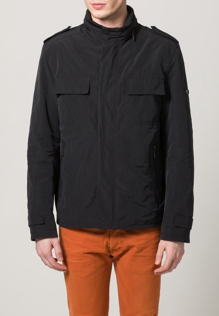 Lekka czarna kurtka z wysokim kołnierzem, dwoma kieszeniami na piersiach, zapinanymi na zamek kieszeniami bocznymi oraz ozdobnymi guzikami. Noszona na pomarańczowych spodniach.