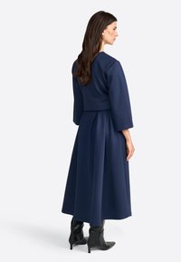 Navy blue, mekko, jossa on pitkät hihat, tiukka vartalo ja laskostettu hame. Valmistettu pehmeästä kankaasta, jossa on pyöreä pääntie ja laskokset vyötäröllä.
