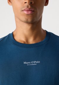 Man met een blauwe ronde hals t-shirt met witte tekst "Marc O’Polo EST. IN STOCKHOLM" op de borst tegen een effen achtergrond.