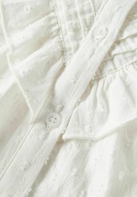 Balta medvilninė palaidinė su tekstūruotu dizainu, puošta raukšlėmis ir apvaliomis balto mygtuko sagomis prie kaklo.
