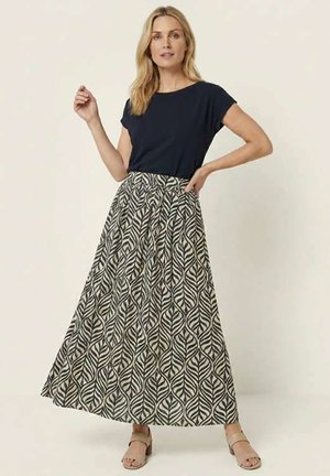 Femme portant un haut noir à manches courtes et une longue jupe beige avec un motif de feuilles noires, debout avec une main sur la hanche.