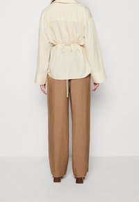 Chemise crème à col, avec un dos cintré, assortie à un pantalon en lin large de couleur marron clair et des chaussures à talons marron. Texture lisse.