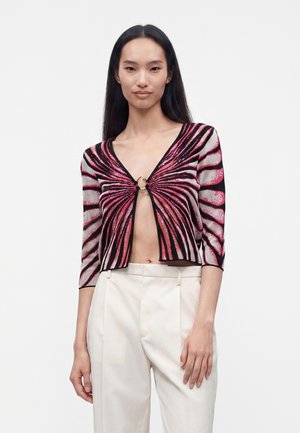 Kvinde iført en lyserød og sort mønstret cropped cardigan med ringelås, kombineret med højtaljede hvide bukser mod en ensfarvet baggrund.