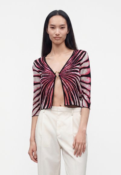 Femme portant un cardigan court rose et noir à motifs, fermé par une boucle en anneau, associé à un pantalon blanc taille haute sur un fond uni.
