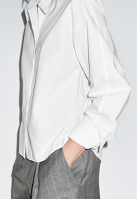 Weißes Baumwollhemd mit lockerem Schnitt, spitzem Kragen und langen Ärmeln. Wird zu grauen, fein gestreiften Hosen mit Seitentaschen getragen.