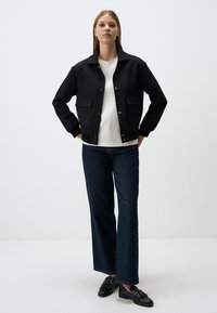 Veste noire à boutons avec col et deux poches avant, superposée sur une chemise blanche. Associée à un pantalon en denim foncé évasé et des chaussures noires.