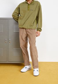 Grön tröja med knappad krage, matchad med ljusbruna kordflortbyxor och vita sneakers med blå detaljer. Mot en gul bakgrund.