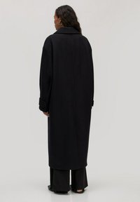 Lange zwarte jas met een gestructureerd ontwerp, grote kraag en oversized mouwen, gemaakt van een textuurstof. Achteraanzicht toont de lengte en silhouet.