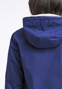 Enfant aux cheveux foncés portant une veste de pluie bleue à capuche, vu de dos sur un fond clair uni.