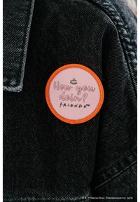 Okrągła naszywka z różową powierzchnią, pomarańczową bordiurą oraz nadrukowanym tekstem „Jak się masz? F.R.I.E.N.D.S.”, z ikoną filiżanki kawy. Przypięta do czarnej kurtki.