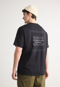 Zwarte katoenen t-shirt met korte mouwen, voorzien van een geprinte witte rechthoekige logo op de achterkant. Draagt het met khaki broek en een crème muts.