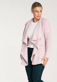 Jasnoróżowy kardigan zapinany z przodu, wykonany z teksturowanej tkaniny, z drapowanym kołnierzem i dwiema przednimi kieszeniami, zestawiony z białą bluzką i ciemnymi dżinsami.