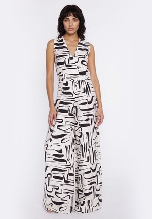 Kobieta w nosząca bezrękawnikowy kombinezon z szerokimi nogawkami w czarno-biały abstrakcyjny wzór, stojąca na jednolitym białym tle.