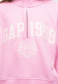 Ροζ φούτερ με λευκό κεντητό κείμενο "GAP 1969" και κυκλικό έμβλημα με καρδιά και στεφάνι δάφνης, με ένα μισό φερμουάρ στον λαιμό.