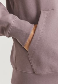 Purple sweatshirt featuring a front pocket, ribbed cuffs, and a smooth texture. Close-up on sleeve and pocket detailing, showing stitching.