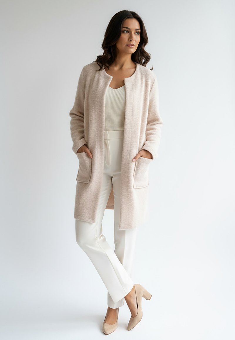 Długi sweter z miękkiej, jasnoróżowej tkaniny z okrągłym dekoltem, dwoma przednimi kieszeniami, zestawiony z białymi spodniami i beżowymi szpilkami.