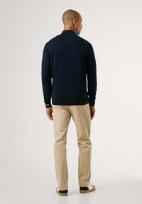 Maglione a collo alto blu navy con finitura testurizzata, maniche lunghe e un piccolo logo sul polsino, abbinato a pantaloni beige e scarpe casual.