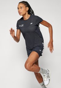 Camisa atlética preta com design texturizado, acompanhada de calções iguais. Apresenta detalhes refletivos e o logótipo da New Balance, usada com meias e sapatos cinzentos.