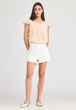 Top color pesco a maniche corte con bordi arricciati, abbinato a pantaloni corti bianchi rifiniti e sandali piatti decorati. Tessuto liscio e design aderente.