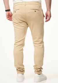 Beige Hosen aus glattem Stoff mit Slim-Fit, ausgestattet mit zwei Gesäßtaschen und braunen Knopf-Akzenten. Getragen mit weißen Sneakers.