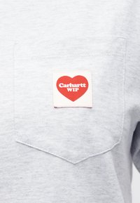 Ανοιχτό γκρι T-shirt με τσέπη, με υφασμάτινο μοτίβο που παρουσιάζει μια κόκκινη καρδιά και το κείμενο "Carhartt WIP". Μαγική υφή, casual σχεδιασμός και μπροστινή τσέπη.