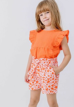 Ein junges Mädchen mit langen Haaren trägt ein oranges, ärmelloses Oberteil mit Rüschen und weiße Shorts mit orangem Herzmuster, steht mit den Händen in den Taschen.