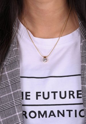 Goldene Halskette mit einem kreisförmigen, klaren Anhänger, getragen über einem weißen Shirt mit schwarzem Textdesign, geschichtet unter einem karierter Blazer.