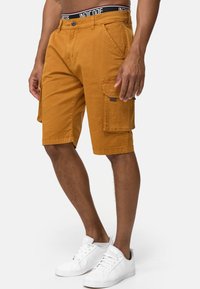 Pantalones cortos de carga color ámbar con una tela texturizada, múltiples bolsillos y cierre de botón. Usados con zapatillas blancas y una cinturilla negra.