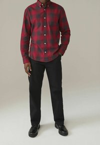 Camisa de franela a cuadros en rojo y negro con cuello abotonado, mangas largas y un bolsillo en el pecho; combinada con pantalones negros y zapatos negros.