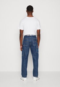 Blå denimbyxor med avslappnad passform, med flera lapplager, sömdetaljer och en kontrasterande varumärkesresår. Bärs med en vit t-shirt.