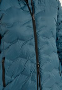 Isoleret dunjakke i dyb teal med kviltet mønster, sorte lynlåse og en glat, blank overflade. Fyldte lommer med lynlås.